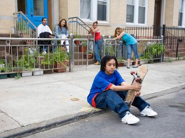 Kid with skateboard sitting on the curb with friends and family in the background