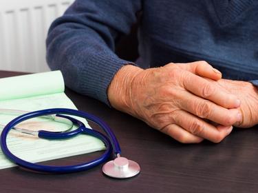 An elderly person's hands on a doctor's desk next to a stethoscope