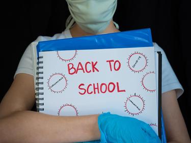 Child wearing a face mask and gloves, holding a binder with Back to School and drawings of coronavirus, photo by Amy Mitchell/Getty Images