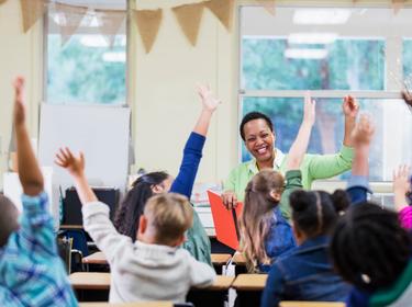 Multiracial group of children raising their hands in a classroom with a smiling Black woman teacher, photo by kali9/Getty Images