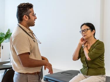 Mature Asian woman talking to a male doctor and indicating her neck and jaw, photo by Fly View Productions/Getty Images